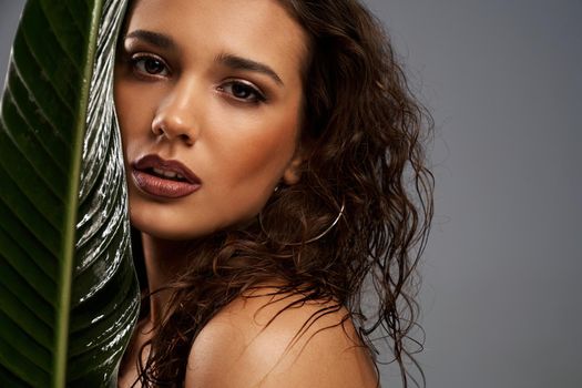 Close up of beautiful naked female model looking at camera. Crop portrait of young brunette woman with perfect makeup posing with mouth open, holding big green leaf, isolated on gray.