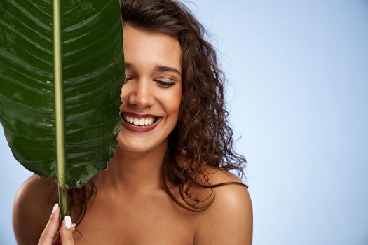 Portrait of young naked woman with perfect makeup laughing and hiding half of face behind green leaf. Close up of beautiful female model posing and looking down isolated on blue.