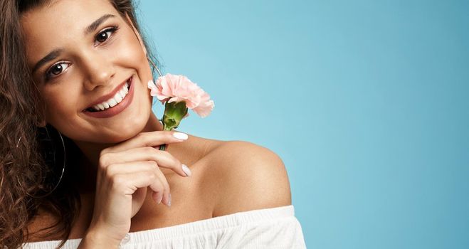 Crop of happy beautiful brunette caucasian female model with curly hair wearing dress with naked shoulders, looking at camera. Pretty woman smiling with teeth, posing holding clove flower near face.