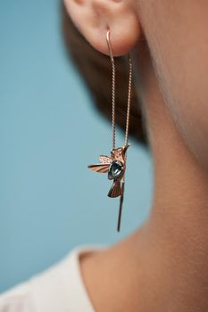 Crop of young female model presenting golden earrings in shape of bird with blue stone. Front view of incognito woman posing, isolated on blue studio background. Concept of jewelry.