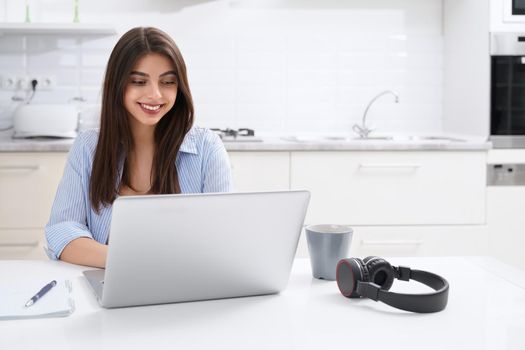 Portrait of beautiful young woman using laptop at home in kitchen. Concept of process studying or working in laptop.