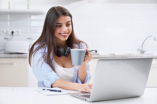 Close up of smiling brunette woman enjoying cup of coffee and studying in laptop. Concept of relaxing and studying at home.