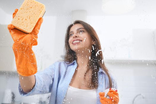 Close up of smiling woman in orange rubber gloves washing window with rag and detergent. Concept of process cleaning.