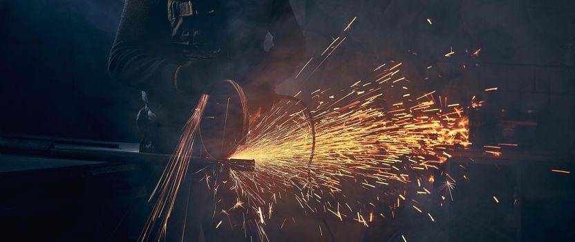 Close up of young man in special dark suit grinding metal on steel pipe with flash sparks. Concept of process working with angle grinder and metal in factory or garage.
