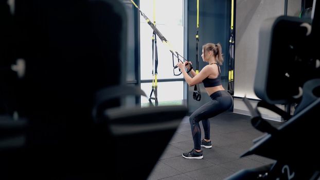 Side view of attractive sporty woman in black sportswear doing exercise and squatting with TRX system. Concept of process workout in modern gym with sports equipments.