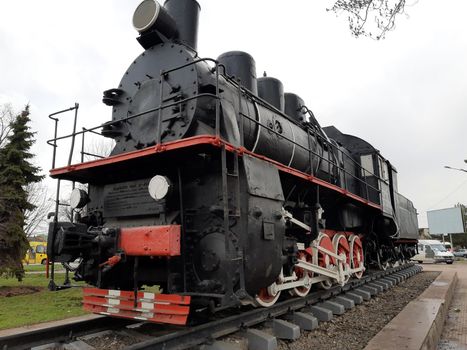 An old black steam locomotive on rails and a concrete platform in the middle of a city square.