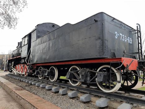 An old black steam locomotive on rails and a concrete platform in the middle of a city square.