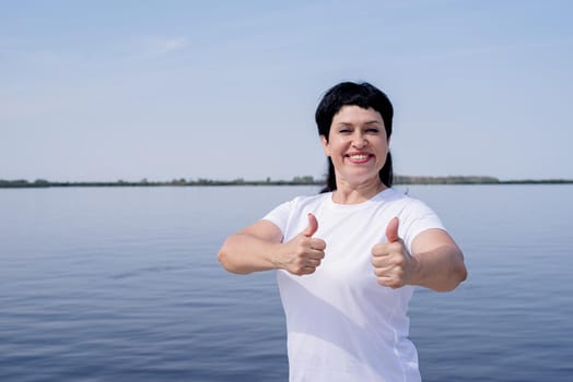 Sport and fitness. Senior sport. Active seniors. Active and happy senior woman in sportswear showing thumbs up working out near the riverside