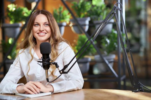 Close up portrait of cute smiling woman in white shirt giving interview by microphone in modern cafe. Concept of process recording video or interview for followers with good mood. 