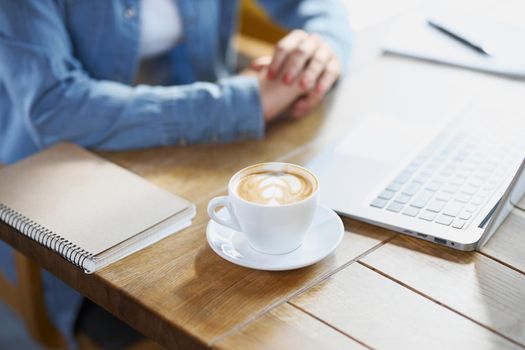 Close up of woman sitting at the table with cup tasty coffee, modern laptop and notebook with pen. Concept of preparing for work in cafe with laptop.