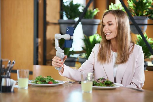 Side view of happy beautiful young blogger woman holding selfie stick with modern phone and doing selfie in cafe. Concept of process communicating online in cafe with tasty food. 