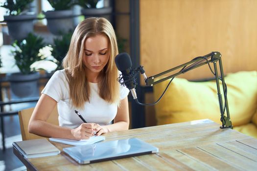 Front view of young attractive woman sitting at the table and writing some information in notebook. Concept of process working with laptop, microphone and notebook, pen. 