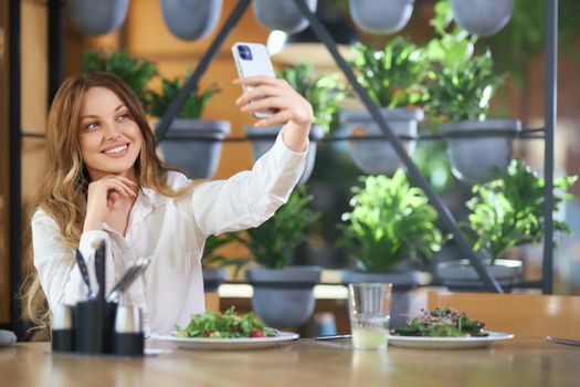 Front view of smiling attractive woman eating tasty food in cafe and doing selfie on modern phone. Concept of process communicating online with friends or followers with good mood. 