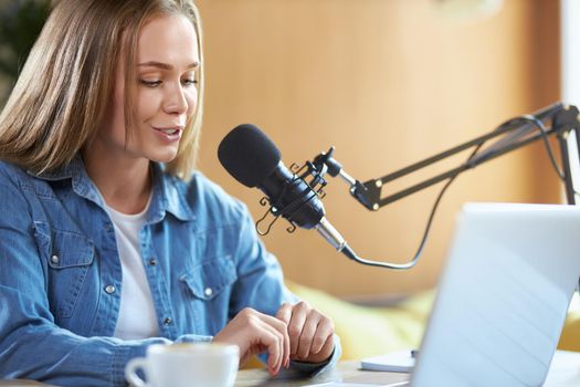 Side view of attractive young woman in denim jacket communicating with people in microphone. Concept of process telling information online with tasty coffee in cafe.