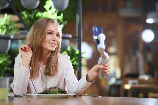 Side view of smiling young attractive blogger woman in elegant jacket sitting in cafe and communicating with followers in live broadcast. Concept of good time with delicious food. 