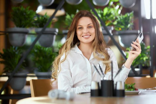 Front view of smiling pretty young woman in white shirt eating delicious salad and chatting with friends on modern phone. Concept of leisure time in cafe with tasty food and communication. 