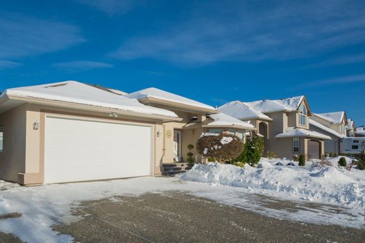 Wide garage of luxury house with driveway and front yard in snow. Street of residential houses on winter sunny day