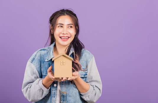 Happy Asian portrait beautiful cute young woman wear denim excited smiling holding house model on hand, studio shot isolated on purple background, broker female hold home real estate insurance