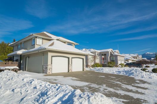 Double garage of big luxury house with driveway and front yard in snow. Street of residential houses on winter sunny day