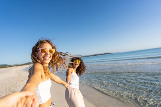 Two young beautiful girls with colorful sunglasses dress boho in summer looking at camera on the beach laughing amused for happiness on vacation pulling hand of the photographer in follow me gesture
