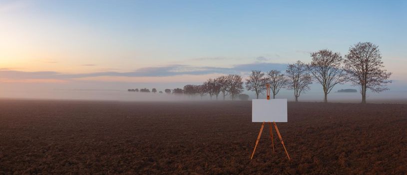 Blank art board and realistic wooden easel on the field. Landscape covered with fog in Central Bohemian Uplands, Czech Republic. Misty morning between fields.