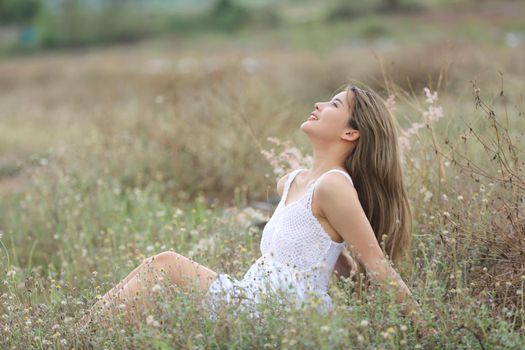 A beautiful woman with long hair in a dress sitting on a meadow and flowers in the evening light.