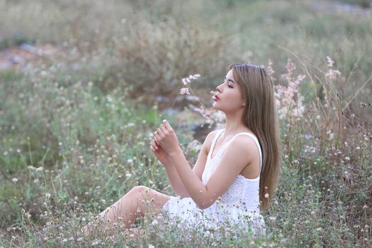 A beautiful woman with long hair in a dress sitting on a meadow and flowers in the evening light.
