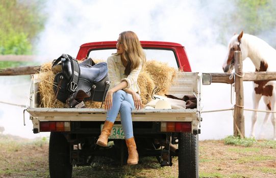 A woman in a cowgirl style sits in a horse ranch with a western farm environment.