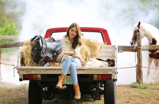 A woman in a cowgirl style sits in a horse ranch with a western farm environment.