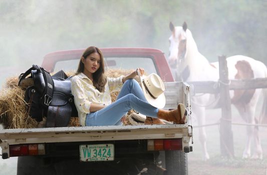 A woman in a cowgirl style sits in a horse ranch with a western farm environment.