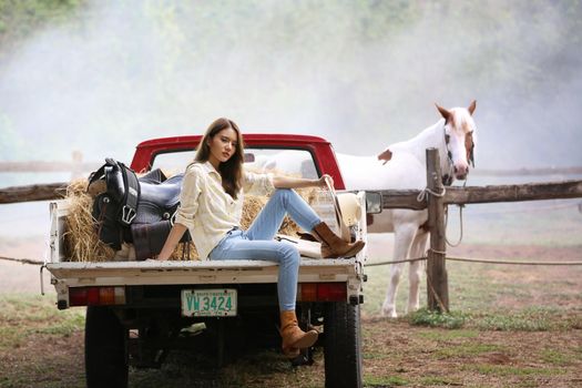 A woman in a cowgirl style sits in a horse ranch with a western farm environment.