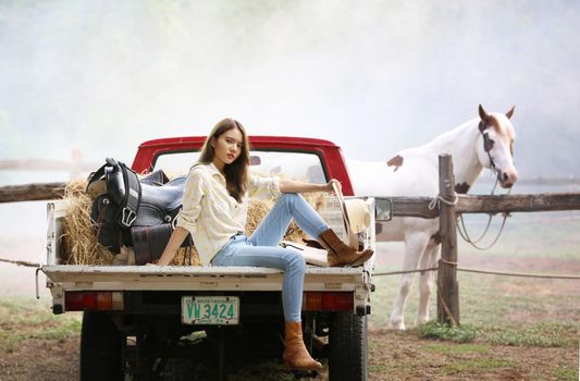 A woman in a cowgirl style sits in a horse ranch with a western farm environment.