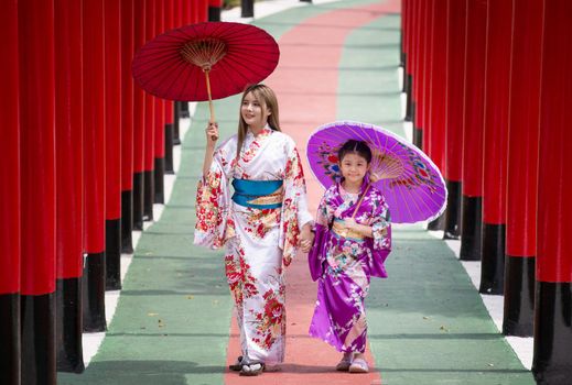  woman and little girl in kimono holding umbrella walking into at the shrine red gate, in Japanese garden.