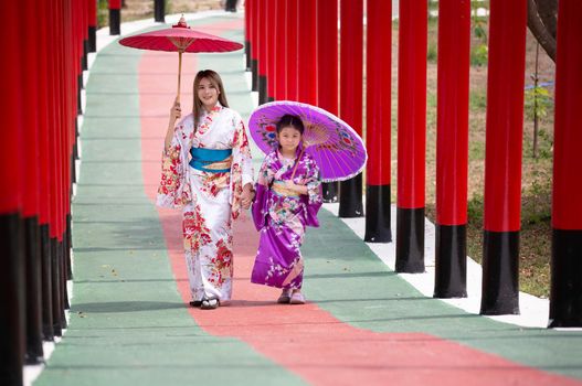  woman and little girl in kimono holding umbrella walking into at the shrine red gate, in Japanese garden.