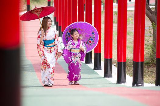  woman and little girl in kimono holding umbrella walking into at the shrine red gate, in Japanese garden.