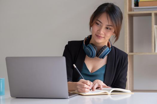 Happy asian woman taking notes in notepad while working at laptop in office. Online Education Concept.