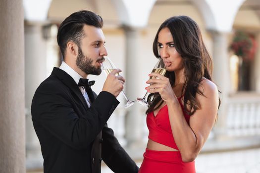 Elegant couple of lovers with a glass of wine or champagne standing on a balcony during luxury party.