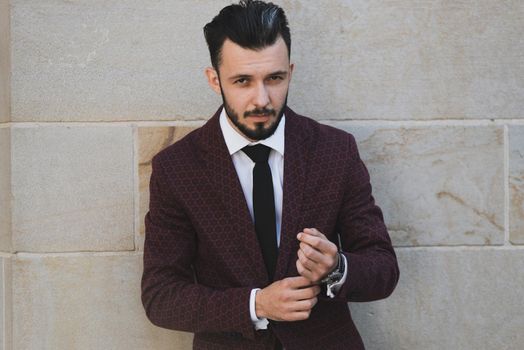 Young and fashionable man buttoning a cuff and posing outdoor on a stone wall background