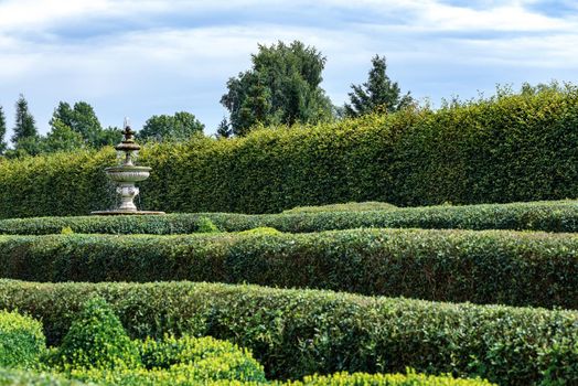 Public garden in a classic style with a green trimmed hedge and a fountain on a sunny day