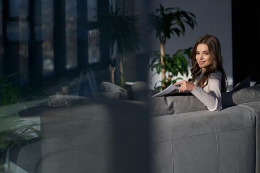 Side view of smiling young brunette beautiful woman sitting on sofa and working or resting with notebook at home. Concept of process working at home with modern interior.