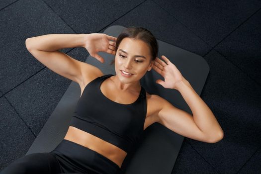 Above view of attractive smiling woman doing abs exercise on black mat in gym. Concept of workout in gym or fitness club for improving body.
