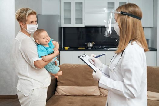 Qualified doctor in medical coat, mask and gloves writing some information on clipboard while young mother standing near and carrying cute baby. Children health care.