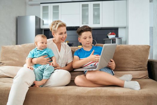Beautiful woman holding on hands her youngest son while sitting on couch near eldest one and using laptop. Mother with two boys playing at home.