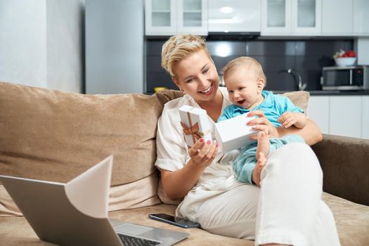 Smiling woman with short blond hair playing with her little son and opening present. Modern laptop and mobile lying on couch near.