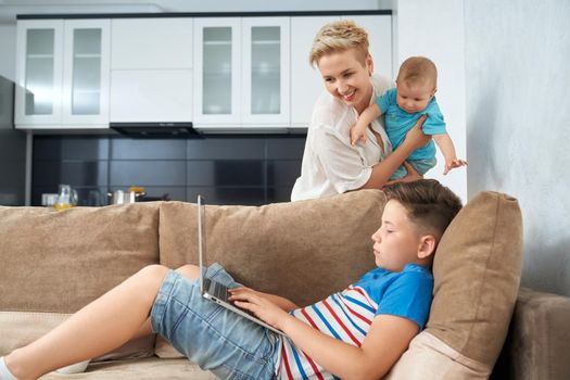 Pretty boy lying on couch with laptop on knees, mother with little baby on hands standing near and looking at computer screen. Concept of family and lifestyle.