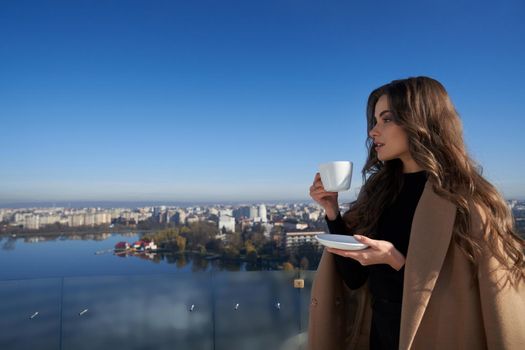 Close up portrait of beautiful young brunette woman standing on balcony with cup coffee or tea and looking on city. Concept of enjoying beautiful nature and city view.