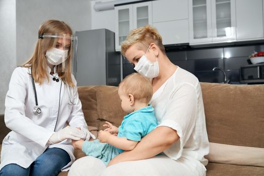 Professional doctor in medical uniform and mask checking health condition of little baby. Young mother holding boy on knees during medical examination.
