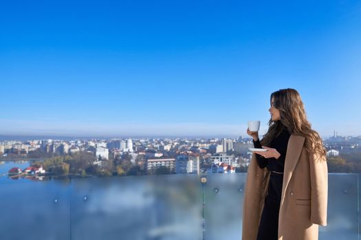 Side view of young beautiful brunette woman in beige coat drinking coffee or tea and looking through panoramic window. Concept of free time with hot drink.