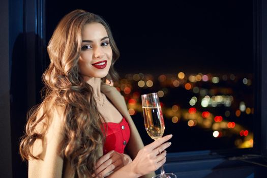 Side view portrait of young beautiful brunette woman standing near window with champagne glasses. Concept of celebration holiday at home with incredible view from window.