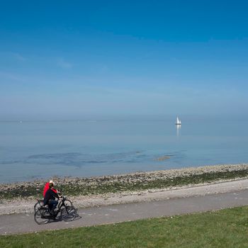 colijnsplaat, netherlands, 31 march 2021: people ride on bicycle track near lonely sailing boat on vast empty blue lake in dutch province of zeeland